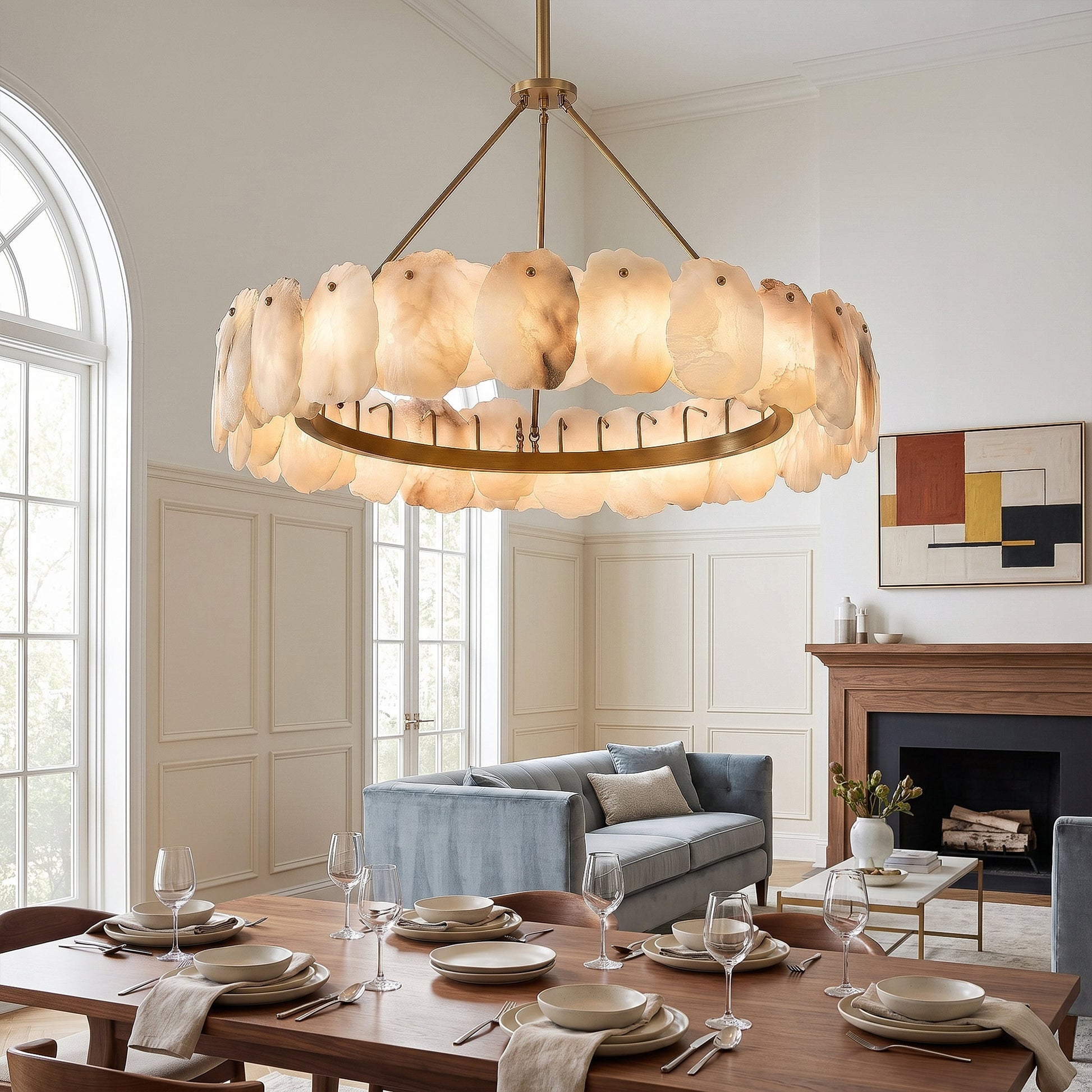 A bright European style open concept dining area and living room with a classic wooden fireplace mantle, a blue velvet sofa, and a large circular alabaster stone pendant chandelier.