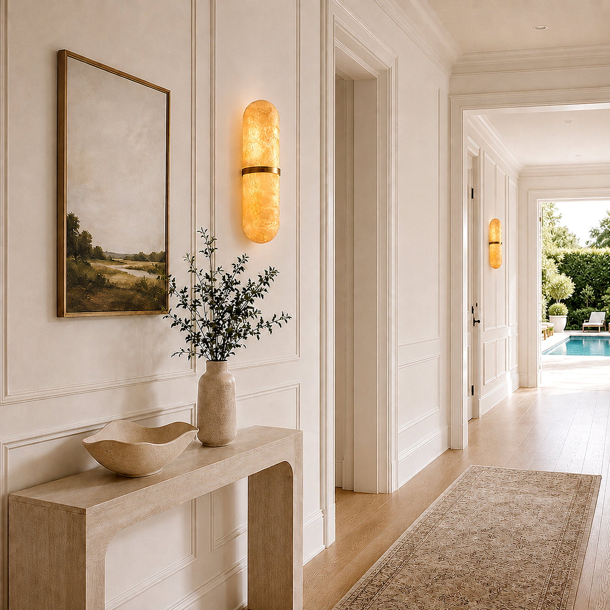 A grand French country estate hallway corridor with light wood floors and classical moldings, featuring a glowing pill-shaped alabaster wall sconce on the right, leading towards an open door with a pool view.