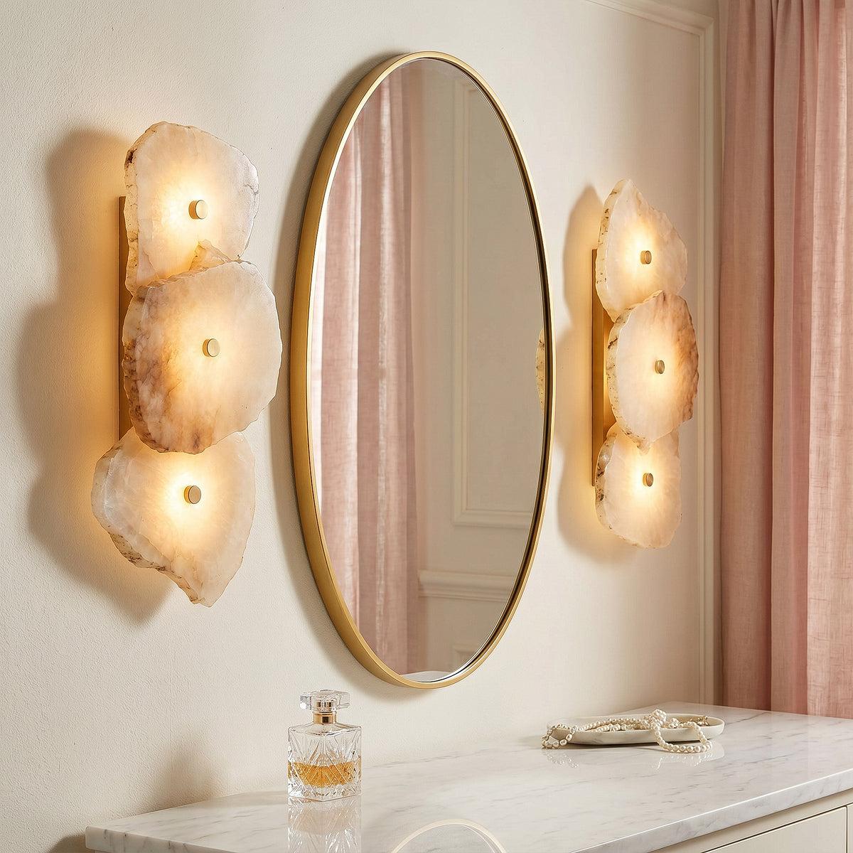 A pair of organic modern Spanish alabaster wall sconces symmetrically framing a gold-rimmed oval mirror over a marble vanity table with pink drapes.