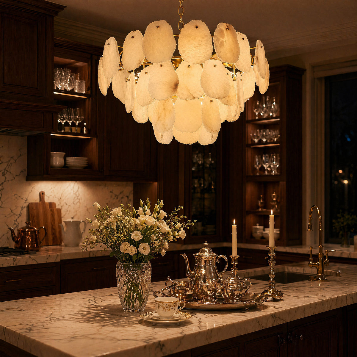A moody upscale kitchen island decorated with tea sets and flowers, highlighted by a large tiered natural stone alabaster chandelier at night.