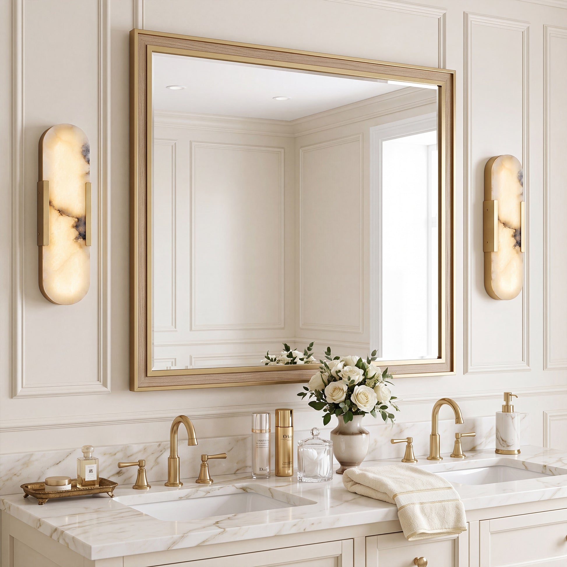 A luxury creamy-toned bathroom double vanity featuring a pair of capsule-shaped Spanish alabaster wall sconces symmetrically framing a large gold-trimmed mirror, set above a white marble countertop with brushed brass faucets and a bouquet of white roses.