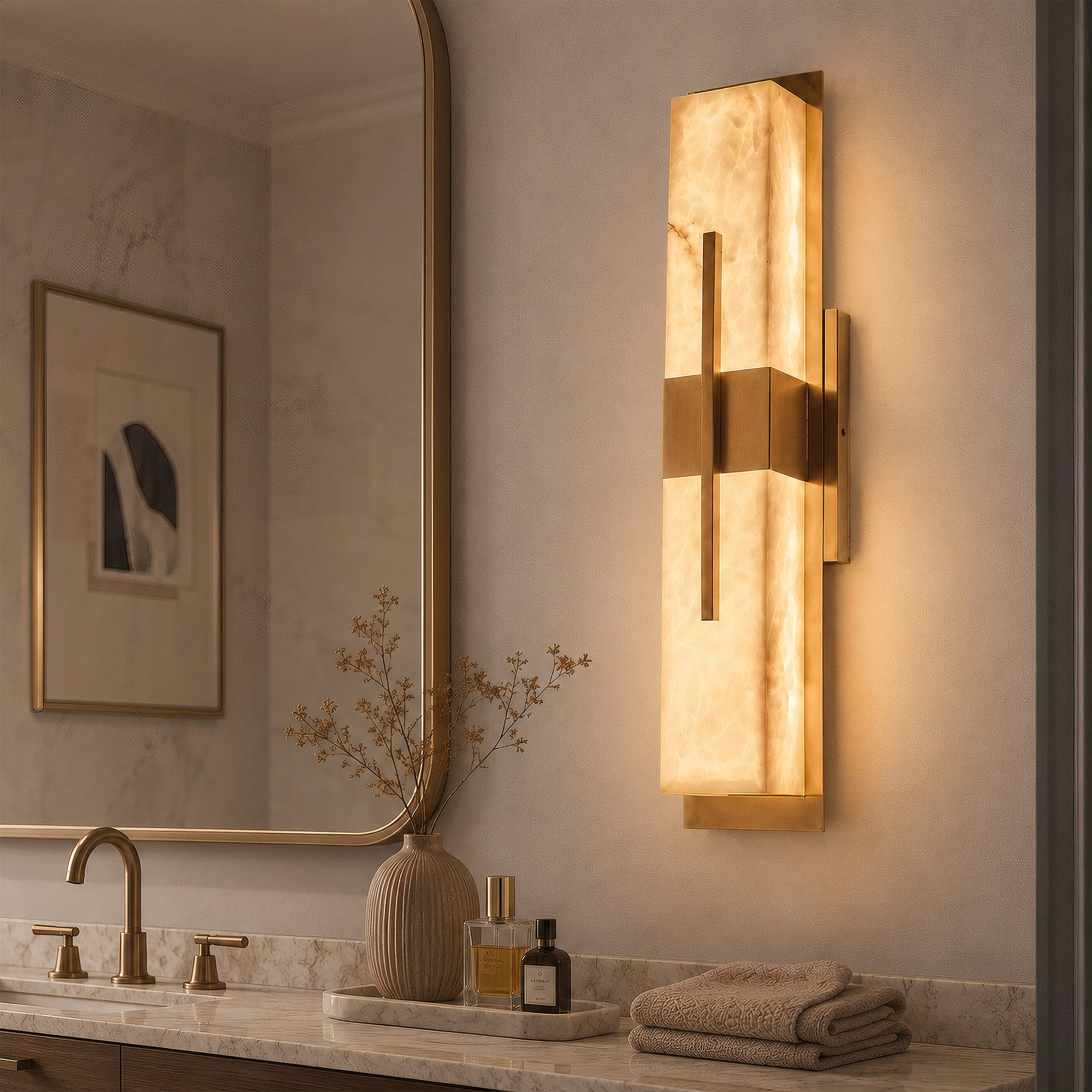 A sophisticated bathroom vanity featuring a vertical geometric brass and alabaster wall light next to a brass faucet, marble countertop, and round mirror reflecting a neutral framed art print.