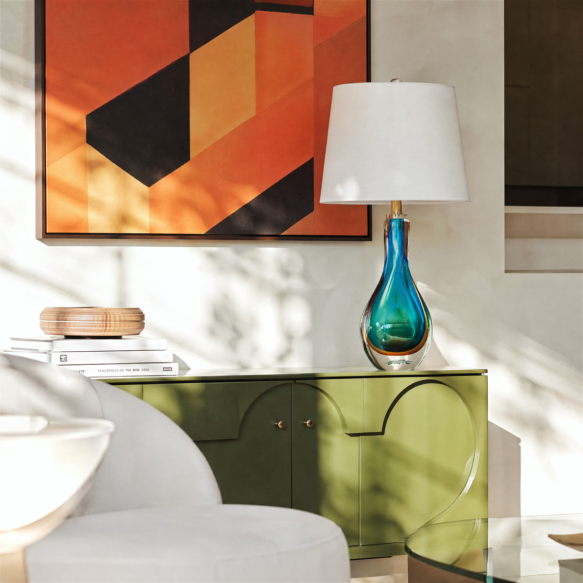 An elegant mid century modern living room setup with shadow play showcasing a teal swirl glass lamp placed on a green sideboard beneath a bold geometric art piece.