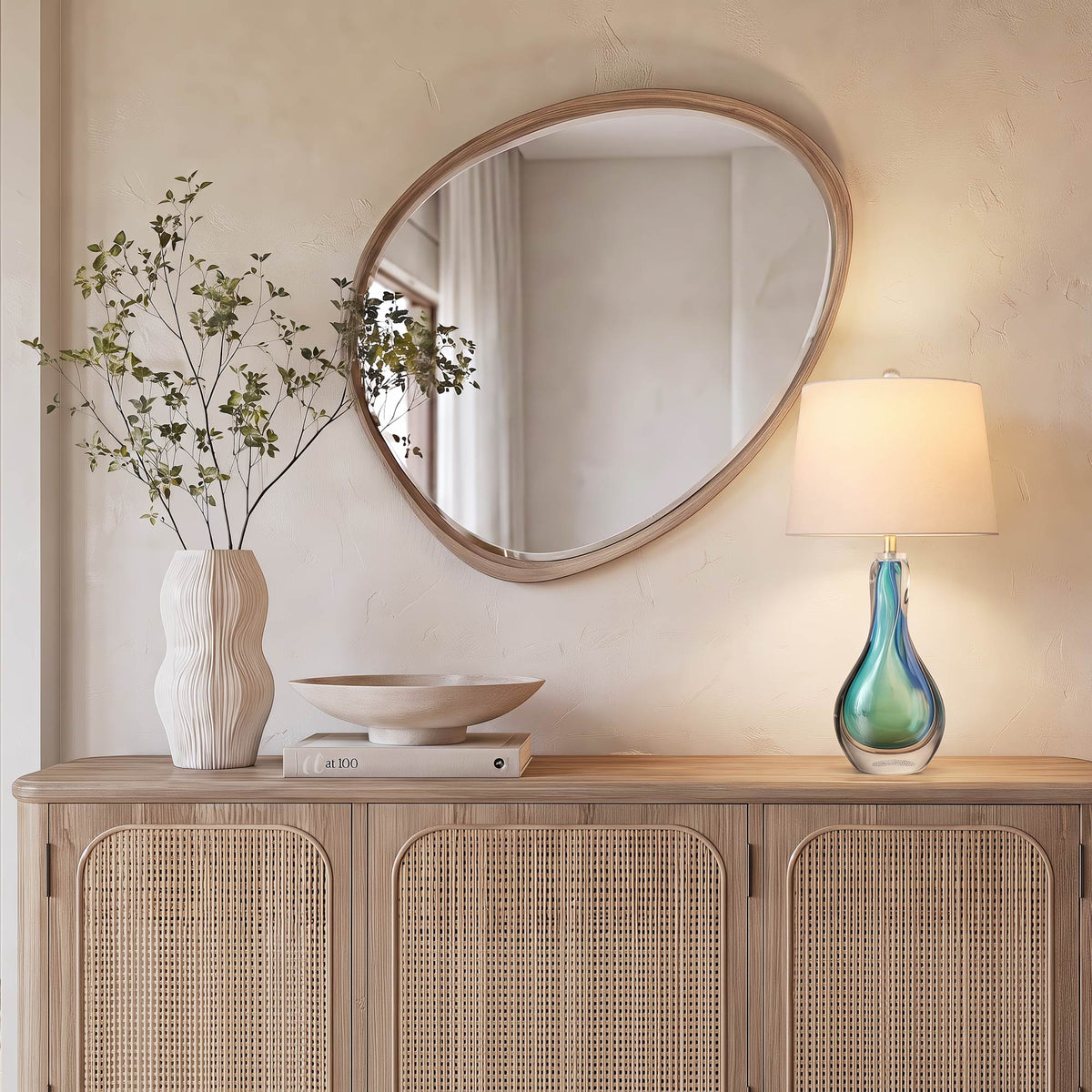 A minimalist cream toned entryway console table featuring a vibrant blue glass table lamp next to an organic wavy white ceramic vase and an asymmetrical wooden mirror.