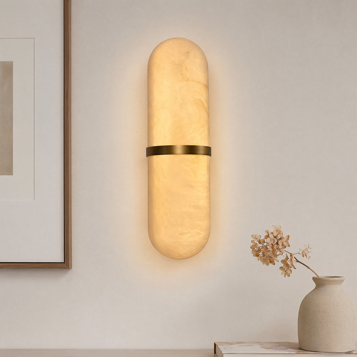 A bright minimalist entryway featuring a glowing pill-shaped brass and alabaster wall sconce on a textured plaster wall next to a large framed art piece and a vase with dry hydrangeas.