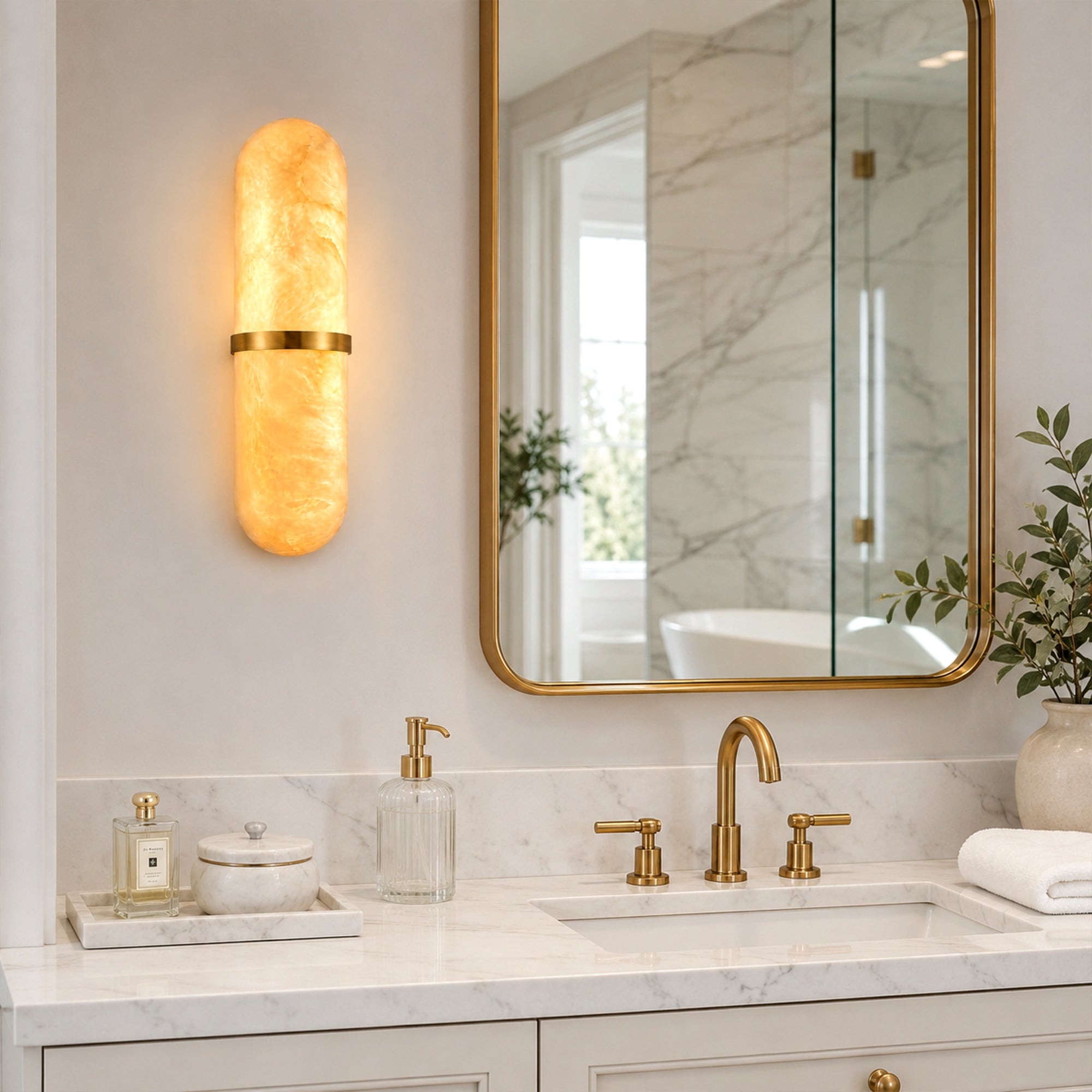 A bright modern bathroom vanity area featuring a pill-shaped Spanish alabaster wall sconce glowing next to a gold-framed mirror, mounted above a single white under-mount sink, white marble countertop, and a brushed brass faucet.