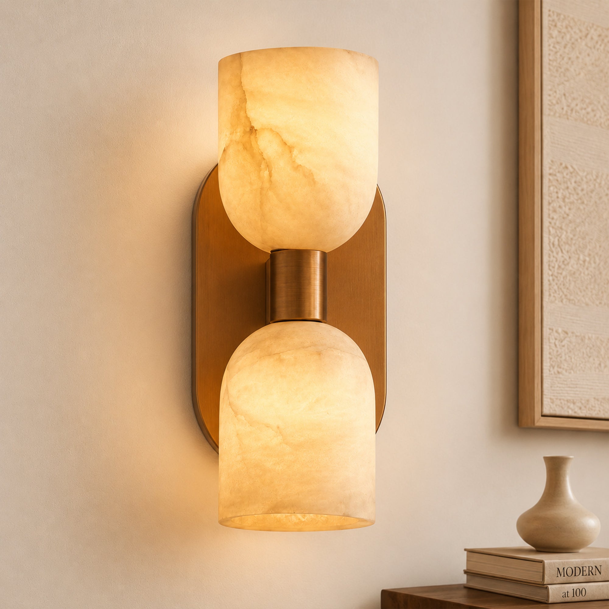 Close-up of a dual-light alabaster wall lamp casting a warm amber glow in an entryway next to a minimalist ceramic vase and books on a wooden console.