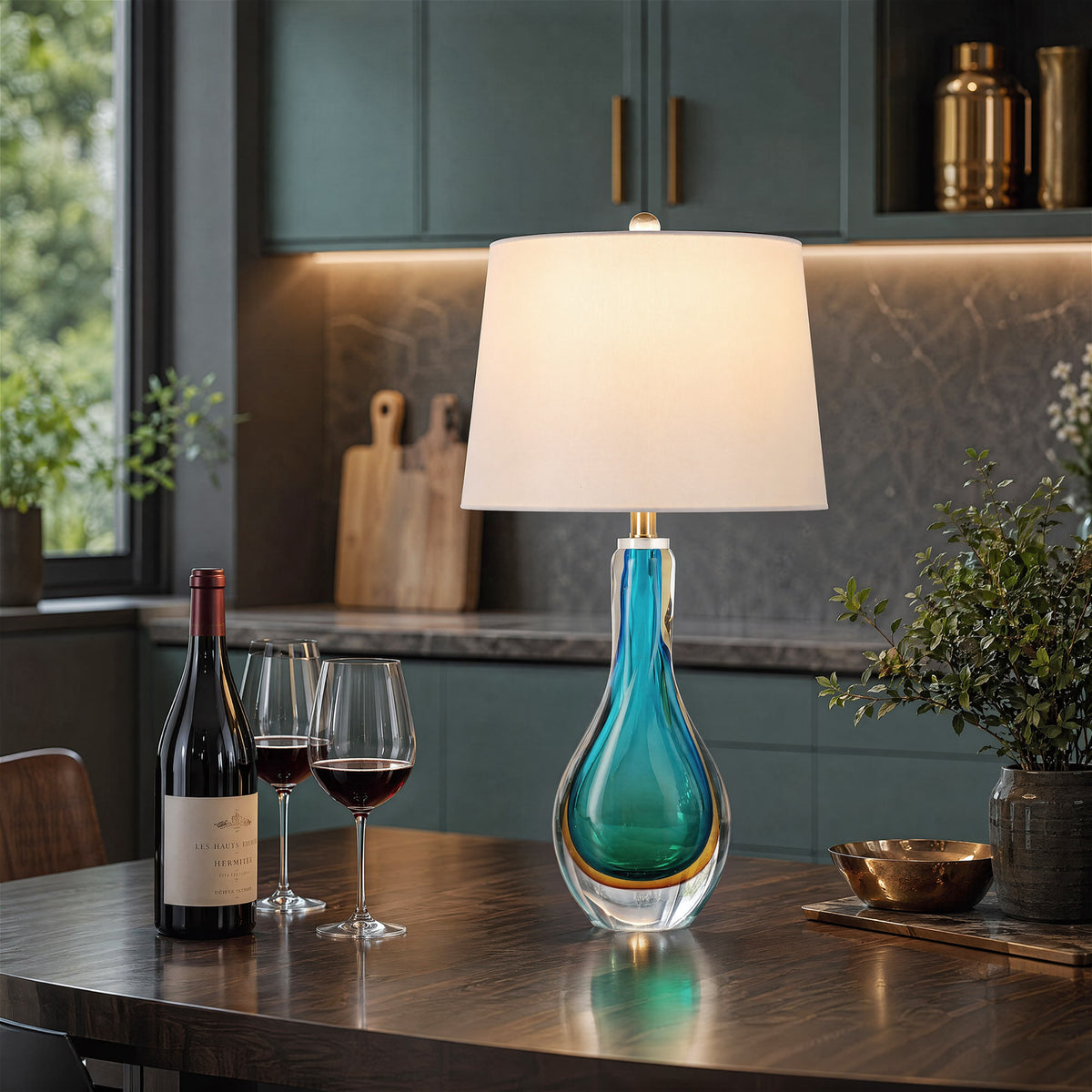 A sleek dark green kitchen dining area highlighting a glowing aqua blue artistic glass lamp on a dark walnut island table accompanied by a bottle of red wine and glasses.