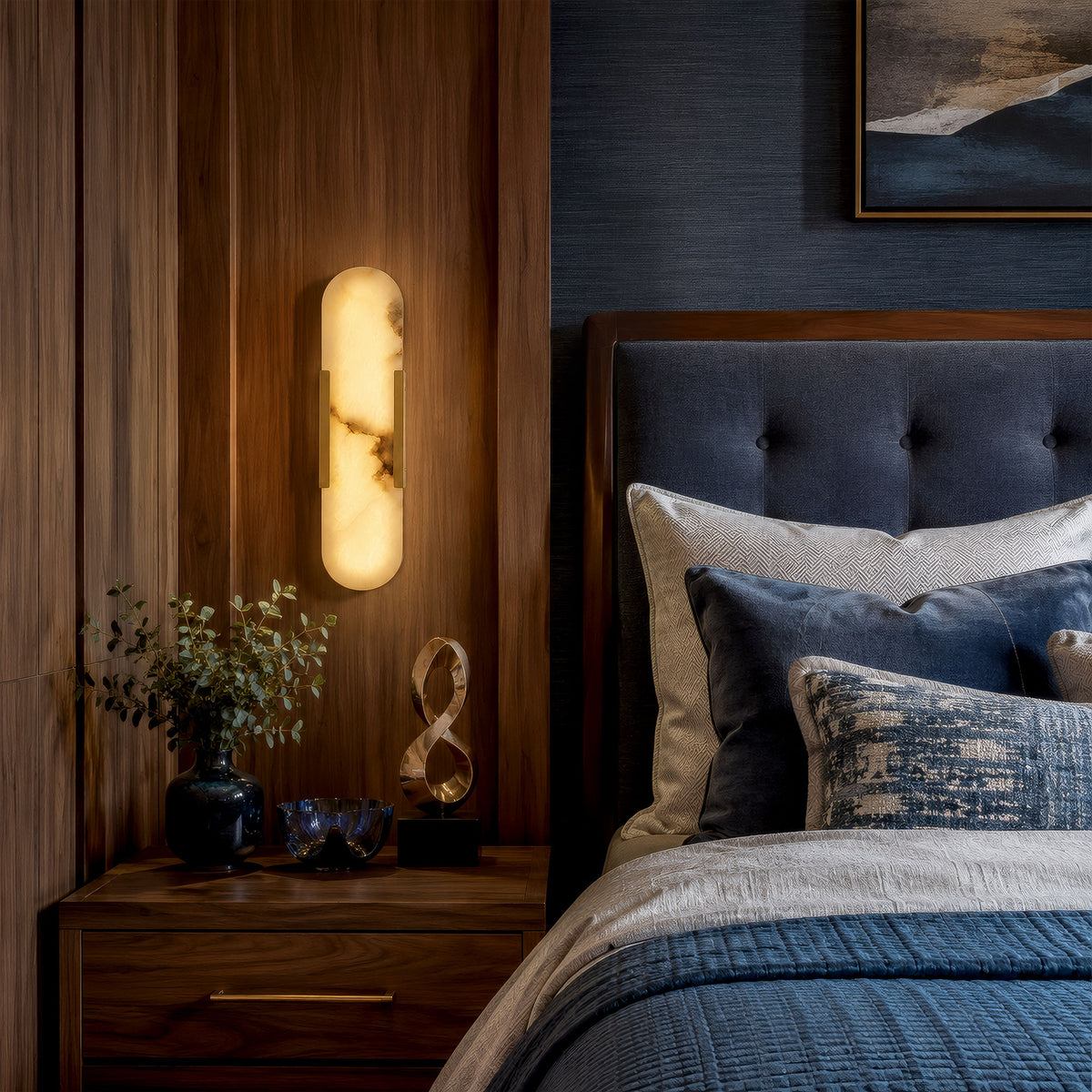 A quiet luxury bedroom setting at night with a walnut wood headboard, a navy blue tufted bed, and a vertical capsule alabaster wall sconce casting a soft golden glow over a nightstand.