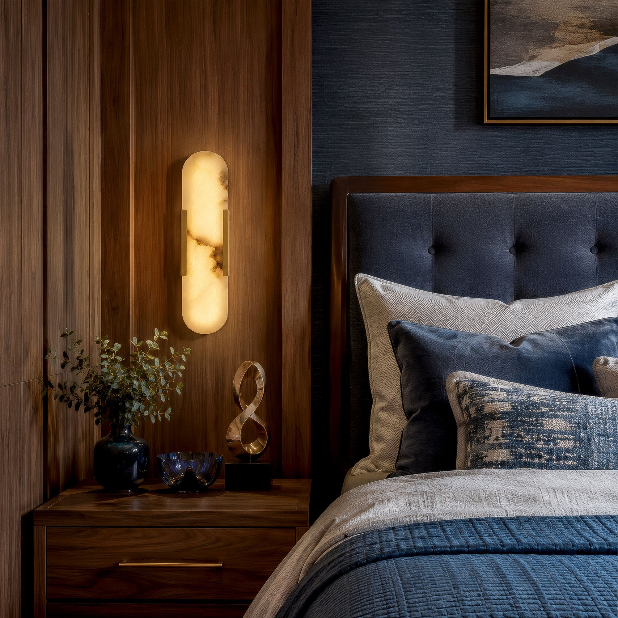 A quiet luxury bedroom setting at night with a walnut wood headboard, a navy blue tufted bed, and a vertical capsule alabaster wall sconce casting a soft golden glow over a nightstand.