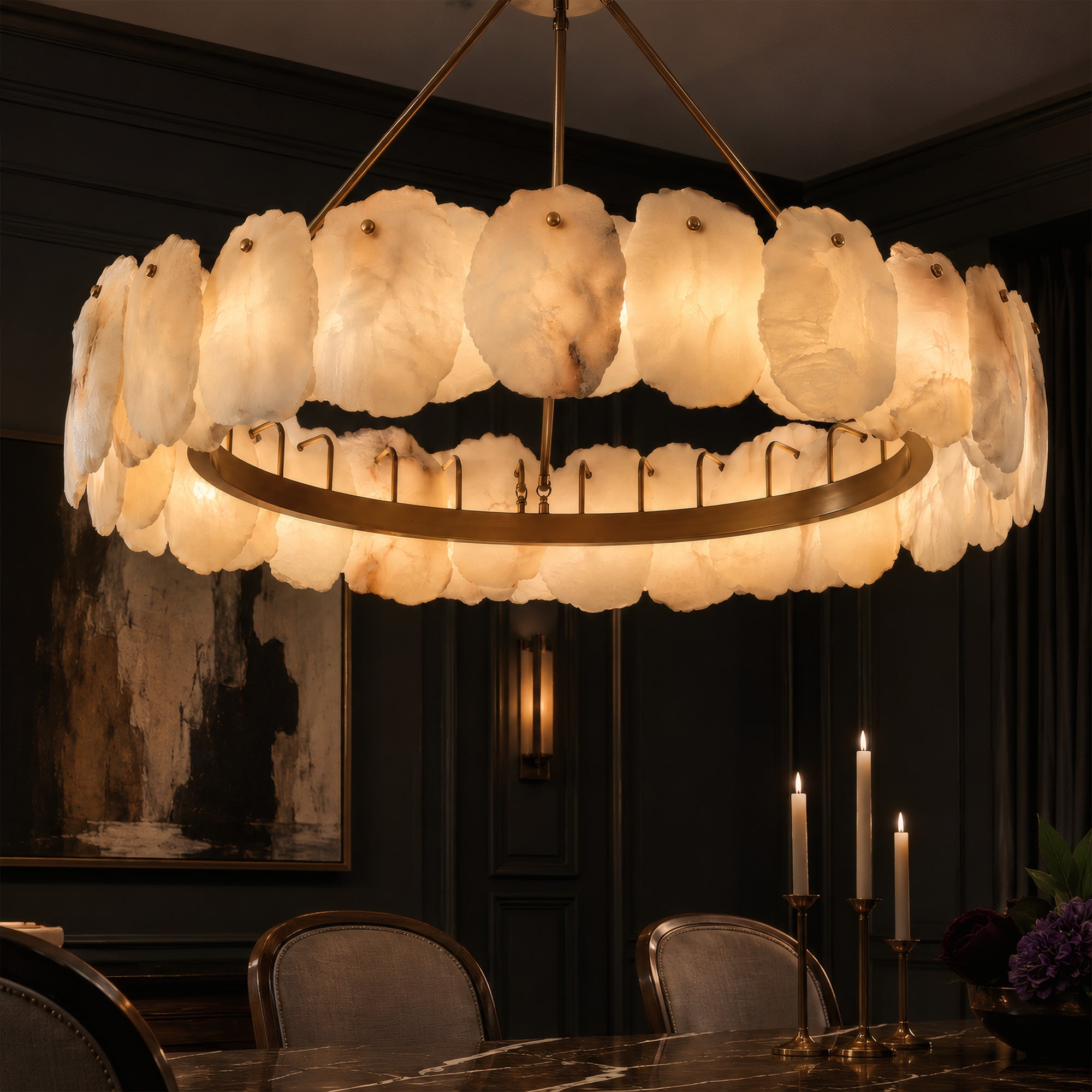 An elegant dark toned luxury dining room at night illuminated by a large round alabaster stone ring chandelier above a black marble table decorated with candles.