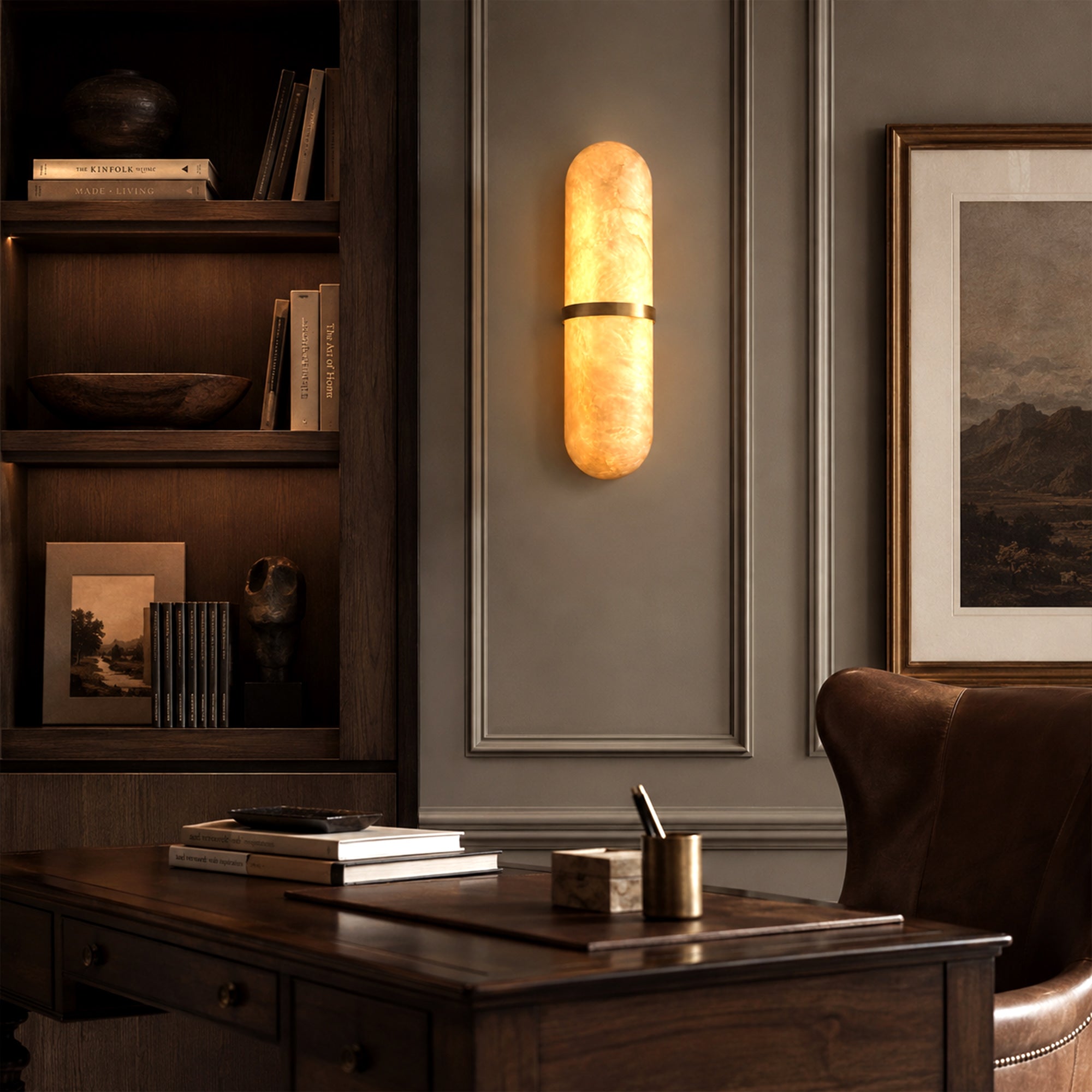 A high-end moody traditional executive library and desk area with dark charcoal-paneled walls, featuring a glowing pill-shaped alabaster sconce mounted next to a dark wood built-in bookshelf and a leather chair.