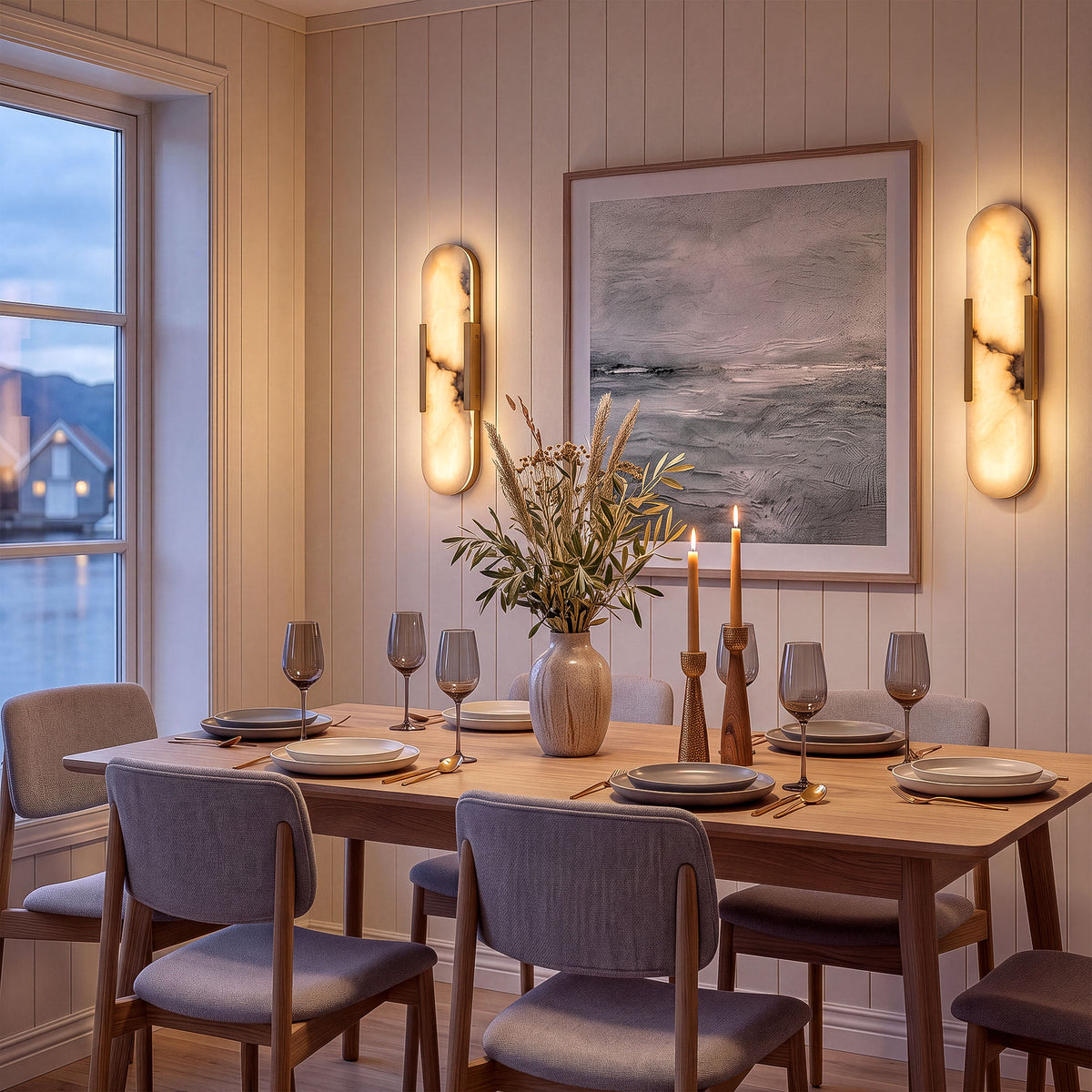 A cozy Nordic minimalist dining room at dusk featuring a pair of oval alabaster wall sconces symmetrically framing an ocean painting on vertical shiplap walls, above a wooden dining table.