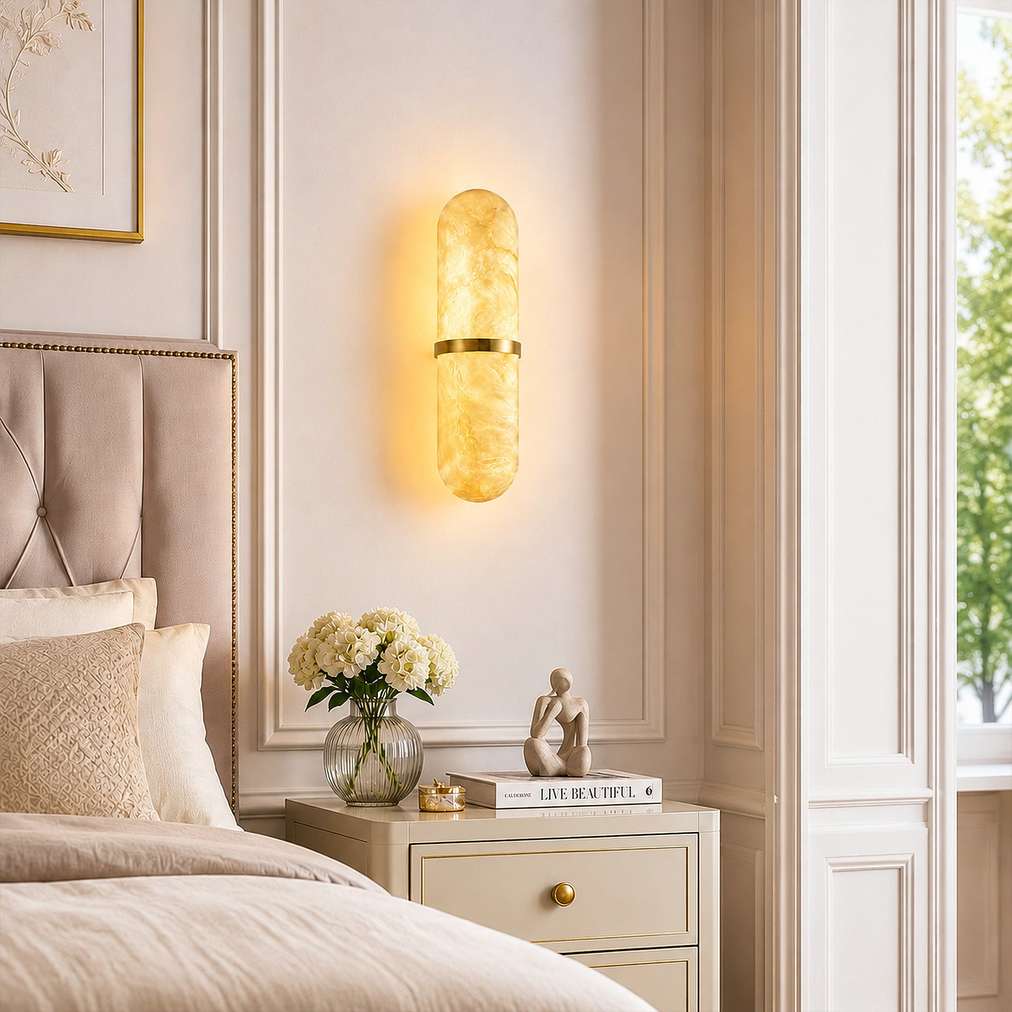 A warm organic modern bedroom corner showcasing a capsule-shaped alabaster wall light mounted on a beige molded wall next to a bedside table set with a bouquet of white flowers and a modern sculpture.