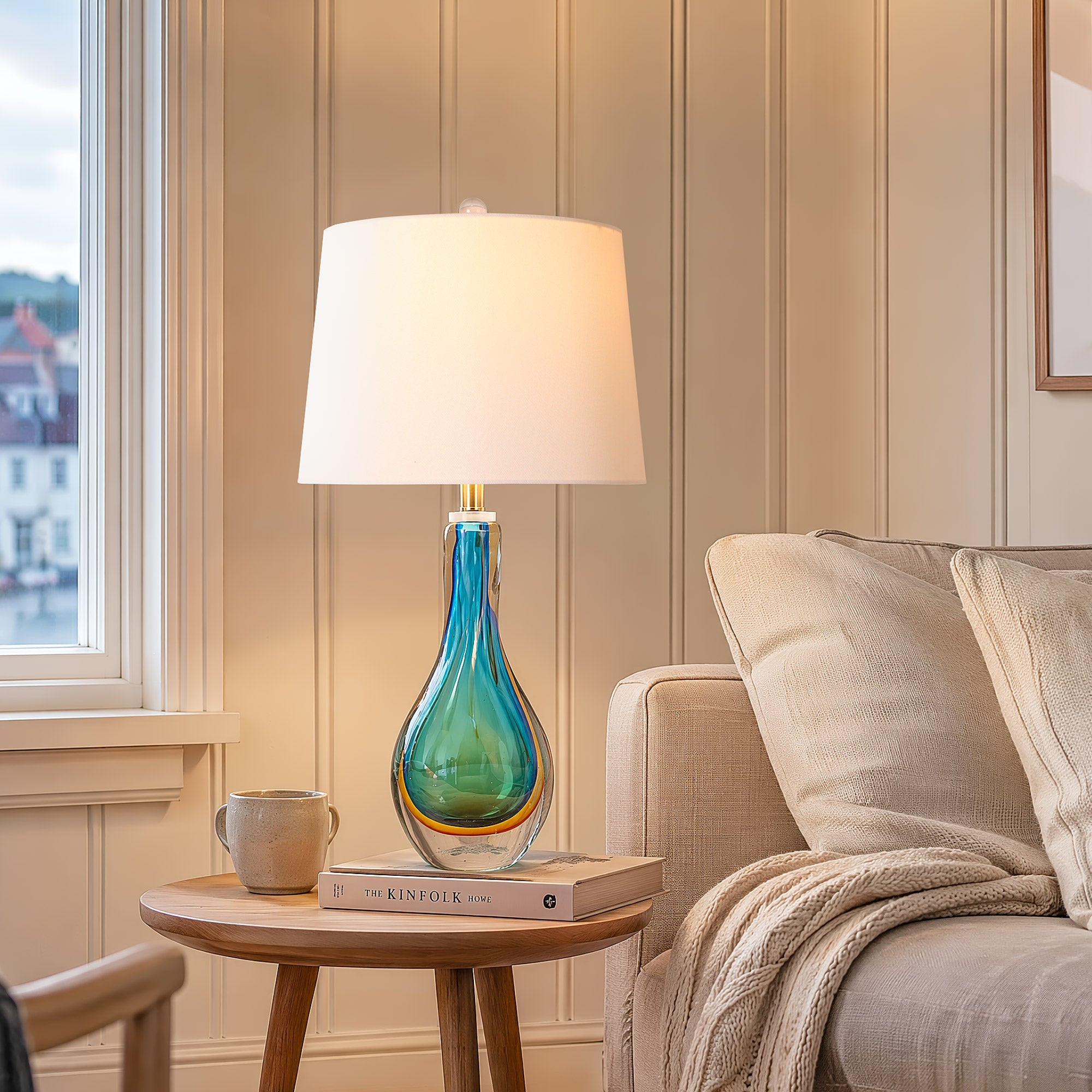 A cozy coastal modern living room scene featuring a lit ocean blue swirled glass art lamp styled on a wooden side table next to a beige fabric sofa with throw pillows.