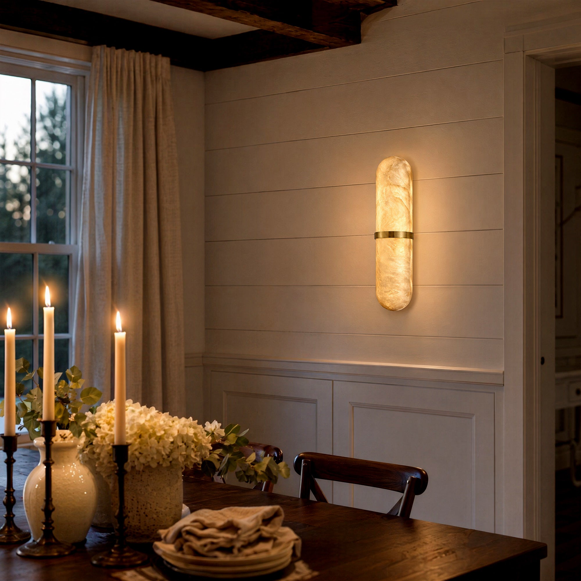 A cozy rustic modern dining room with horizontal white shiplap walls and exposed beams, featuring a glowing brass and alabaster wall lamp casting a soft amber light over a dining table set with white roses and burning candles.