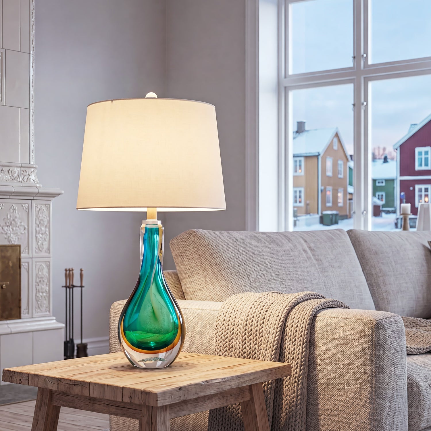 A bright Nordic style living room showing a glowing blue teal art glass lamp on a rustic wood coffee table next to a textured sofa and a classic white tiled fireplace.