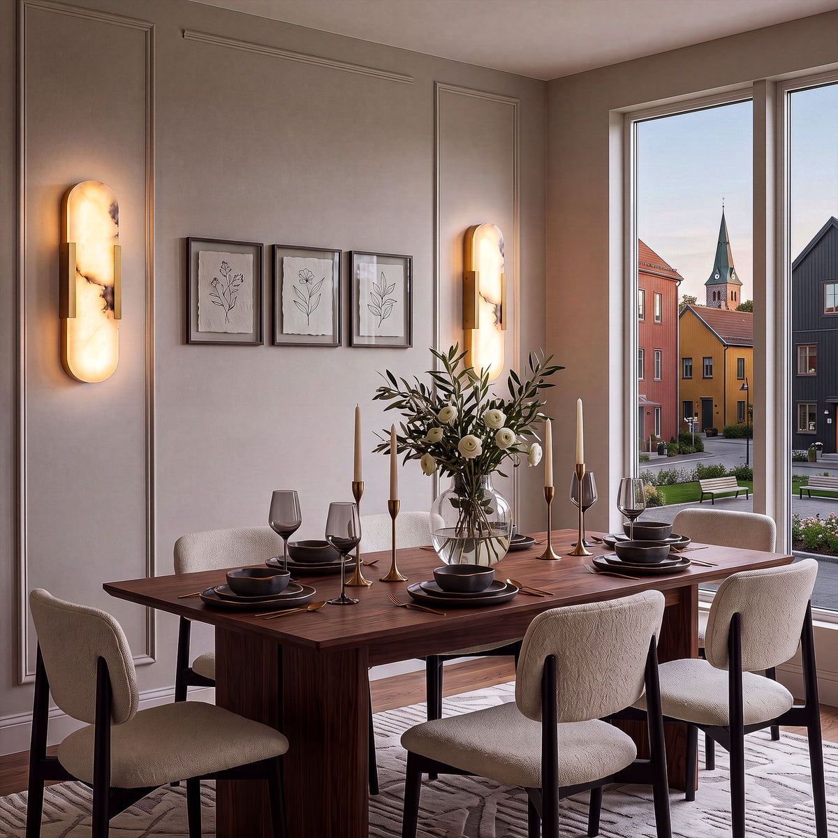 A sophisticated warm taupe dining space featuring a pair of glowing oval alabaster wall lights flanking minimal floral prints, next to a large window overlooking a European village street.