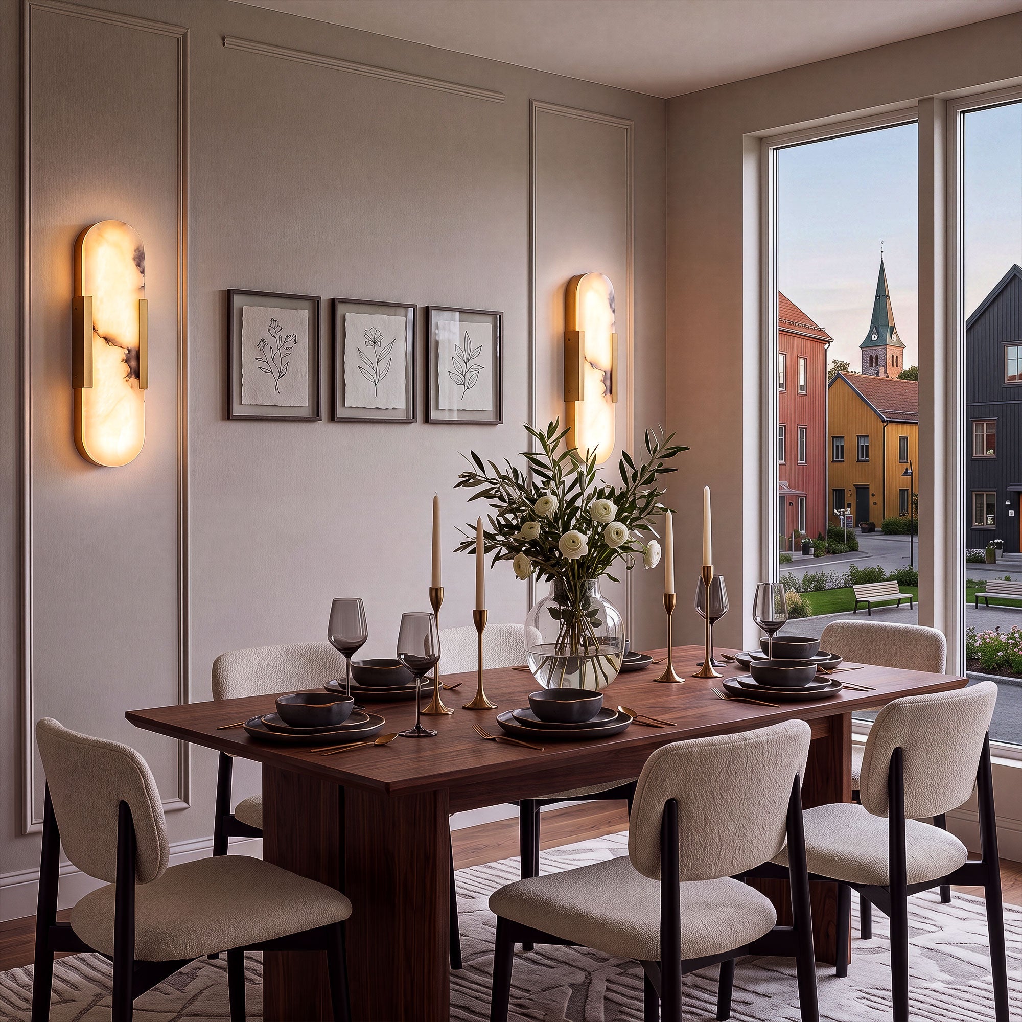 A sophisticated warm taupe dining space featuring a pair of glowing oval alabaster wall lights flanking minimal floral prints, next to a large window overlooking a European village street.