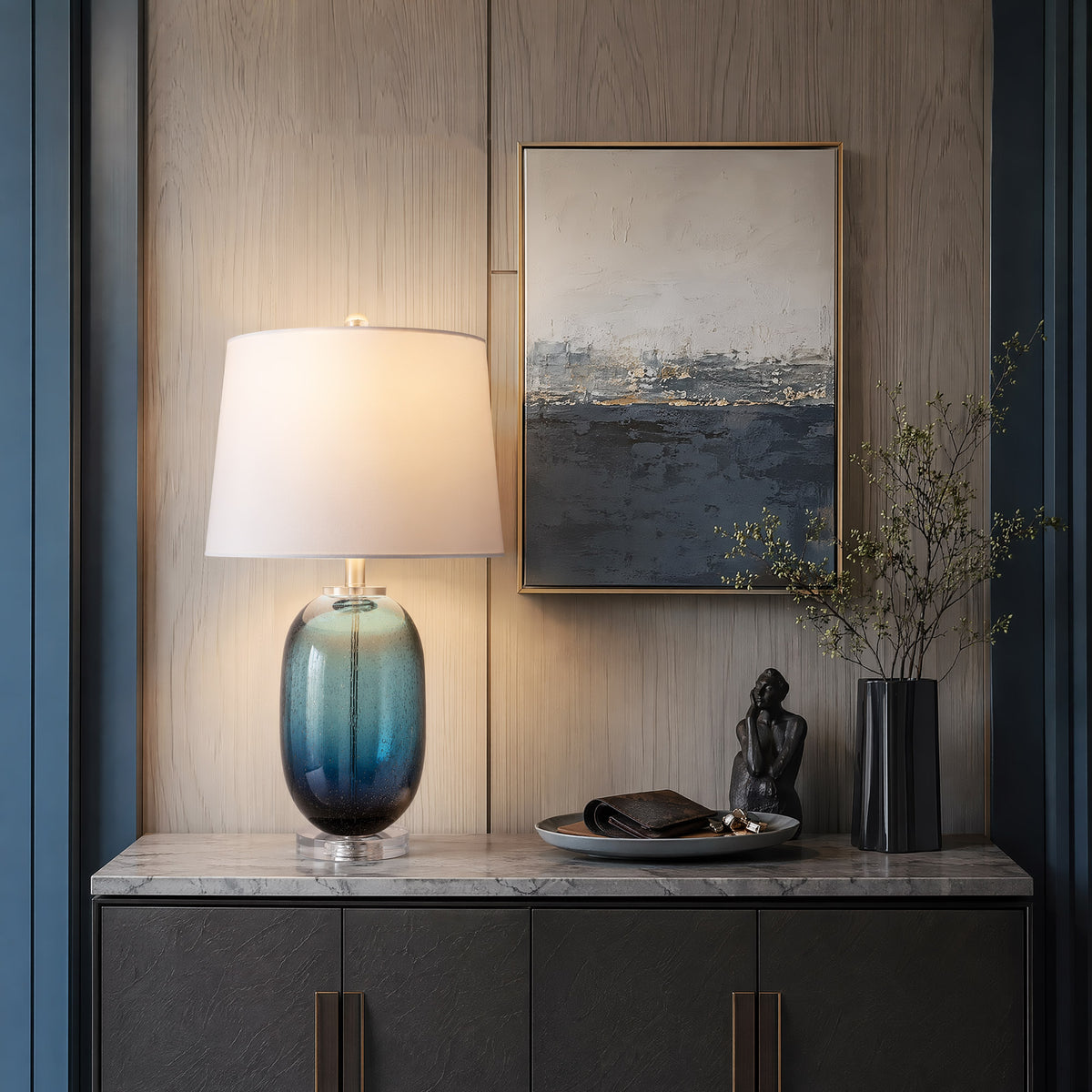 Luxury entryway console table decorated with a glowing tall barrel blue glass table lamp with internal floating bubbles alongside modern abstract seaside wall art