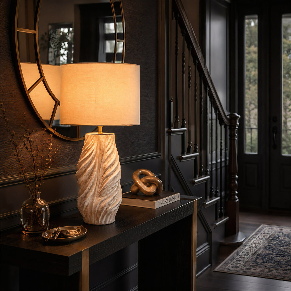 An elegant entryway console table styled with a lit twisted ceramic lamp under a round mirror next to a dark wood staircase