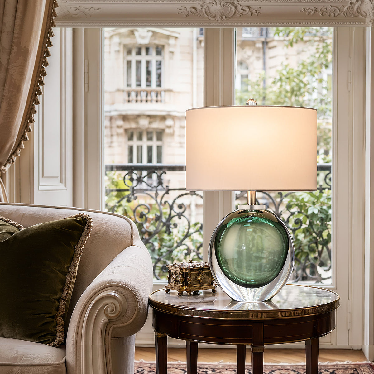 Premium emerald green bubble glass table lamp illuminated on a vintage marble top side table next to a classic cream sofa by a Parisian balcony window