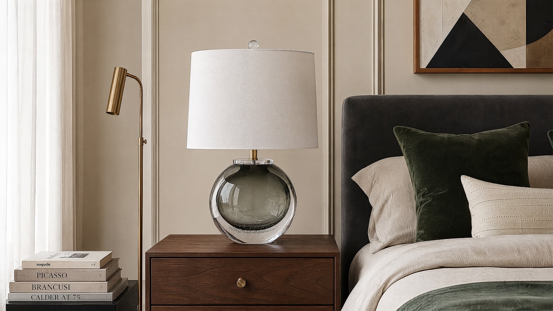 Smoke gray art glass table lamp with white drum shade on a wooden nightstand beside a cozy bed