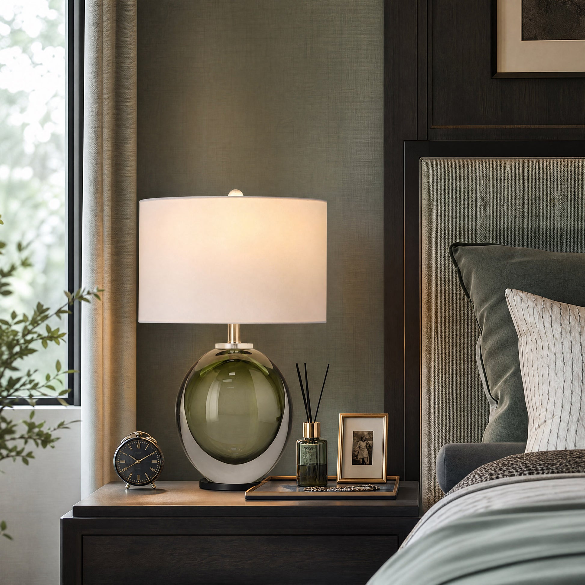 Front view of dark green art glass table lamp with white fabric shade on a dark wooden nightstand in luxury bedroom.