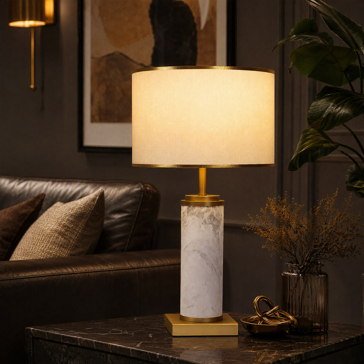 Illuminated white marble cylinder table lamp on dark marble side table beside leather sofa