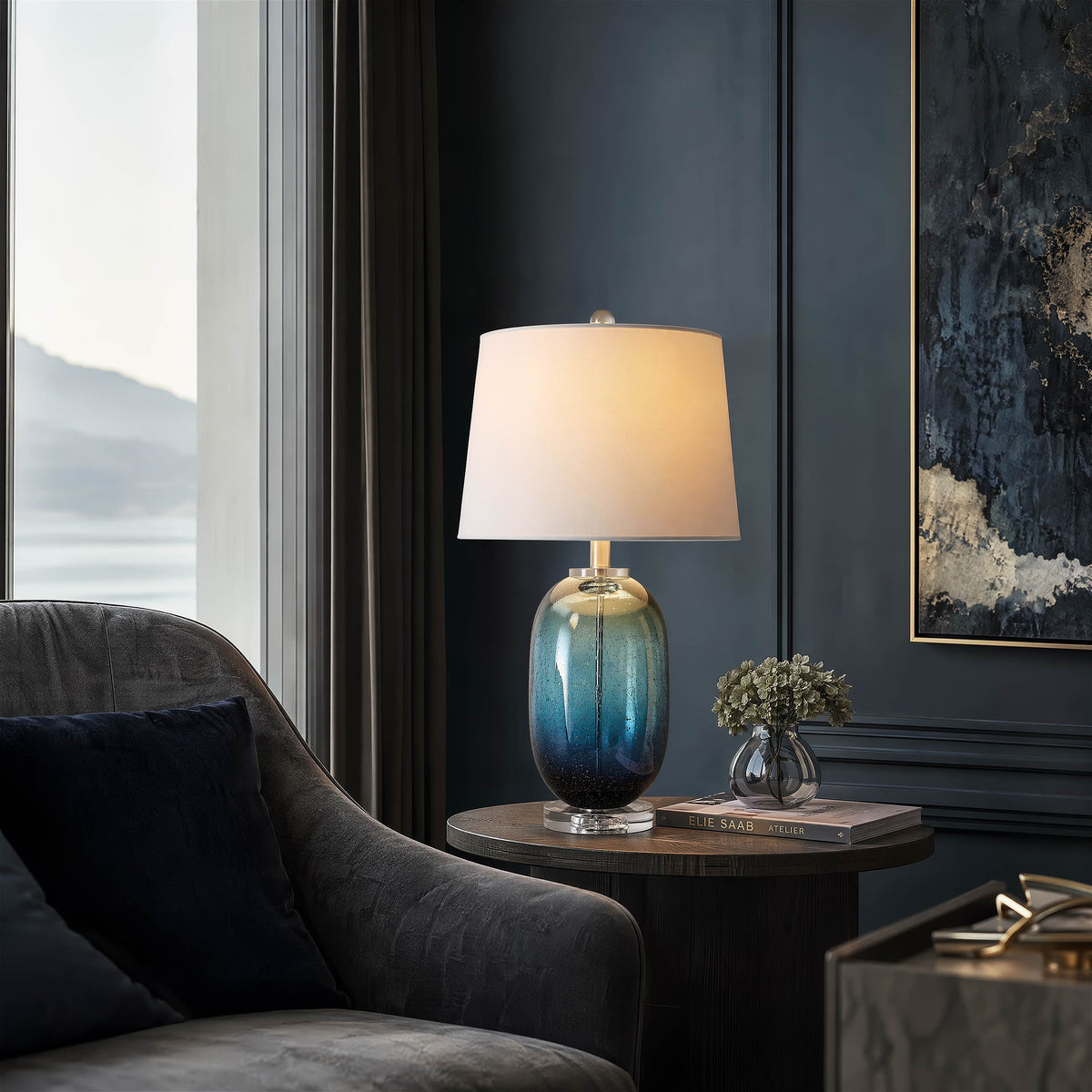 Coastal inspired tall barrel blue art glass table lamp casting a warm light on a wooden accent table near an armchair in a sunlit living room