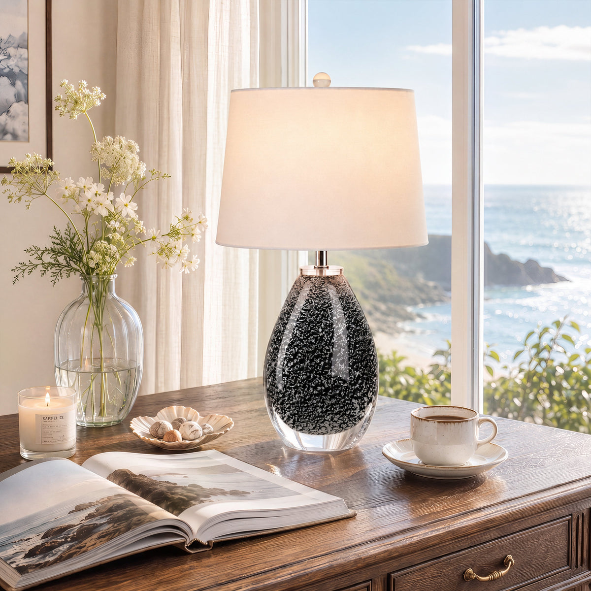 Illuminated glass table lamp on wooden study desk near big window overlooking ocean view.