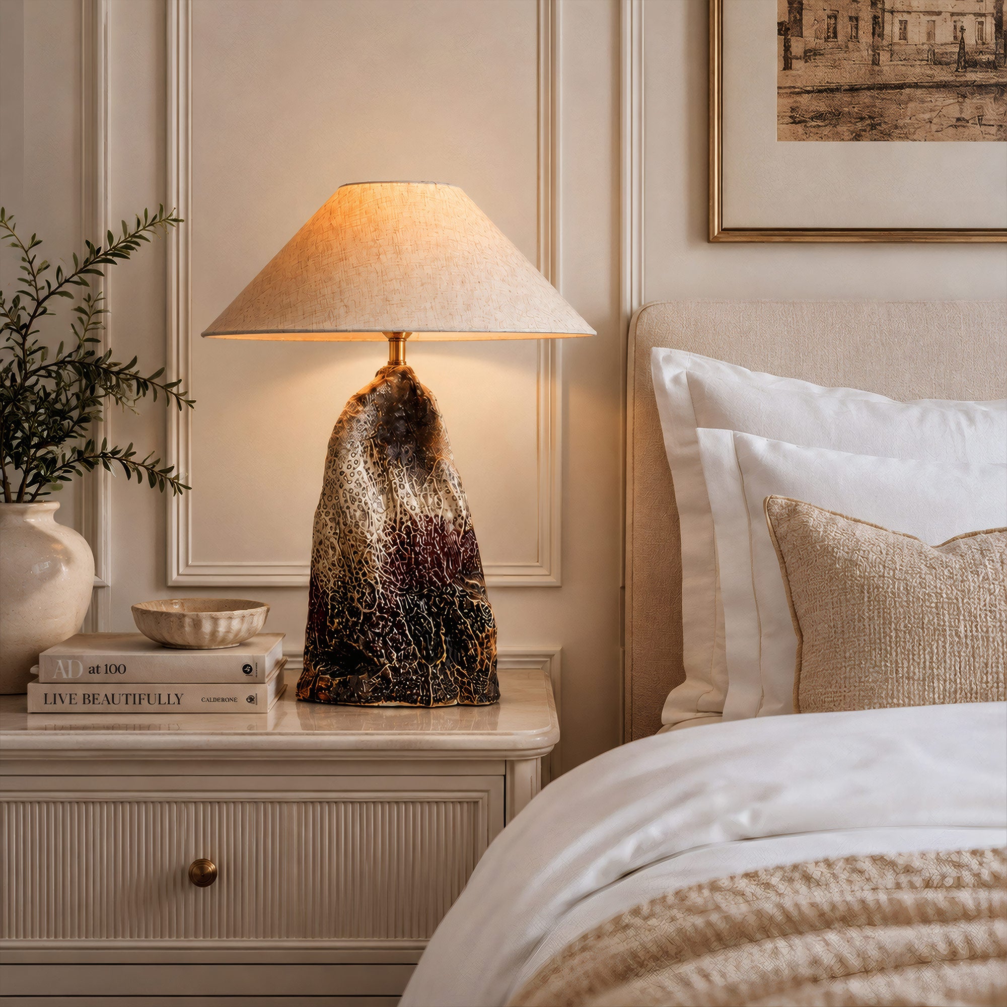 Illuminated rockery flambe glaze ceramic table lamp on a white nightstand bedside table next to a cozy bed in a luxury cream tone bedroom.