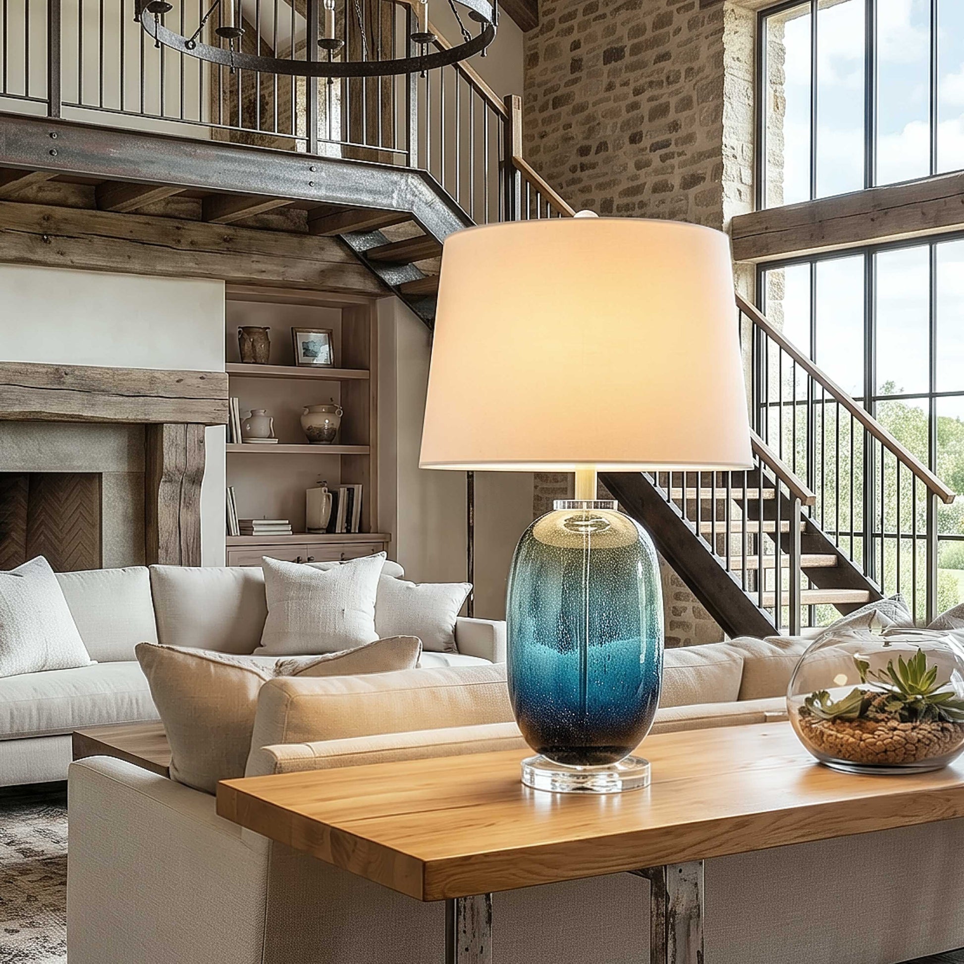 Artistic tall barrel blue seeded glass table lamp lit up on a rustic wooden console table behind a white linen sofa in an airy open layout house