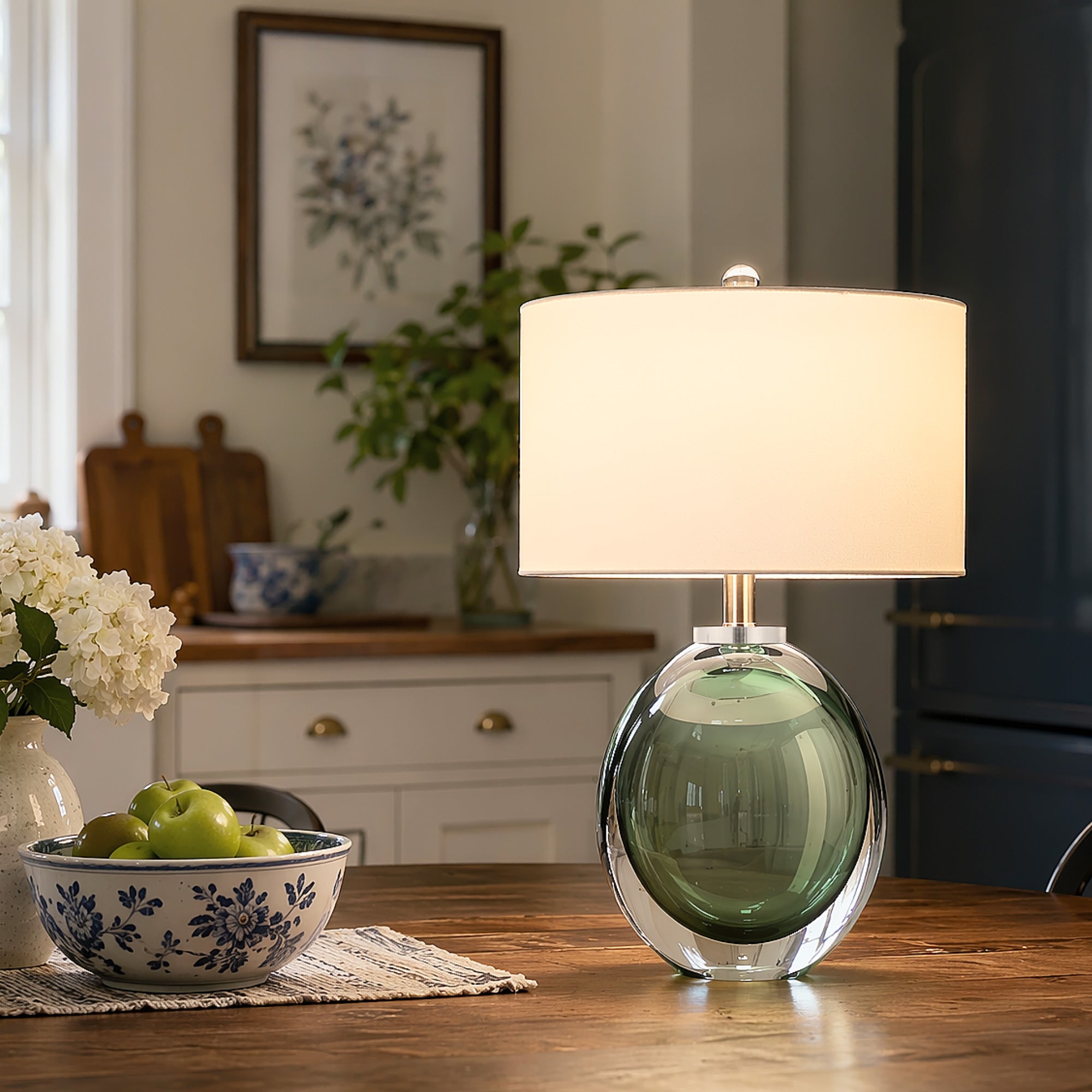 Close up view of an illuminated moss green seeded glass table lamp on a rustic wooden dining table next to a ceramic bowl of green apples in a cottage kitchen