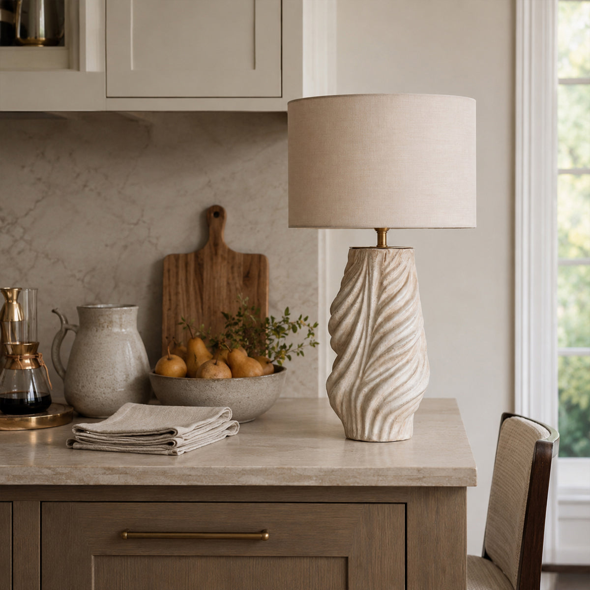 A rustic modern kitchen interior featuring a sculptural fluted ceramic table lamp on a marble countertop next to a bowl of pears