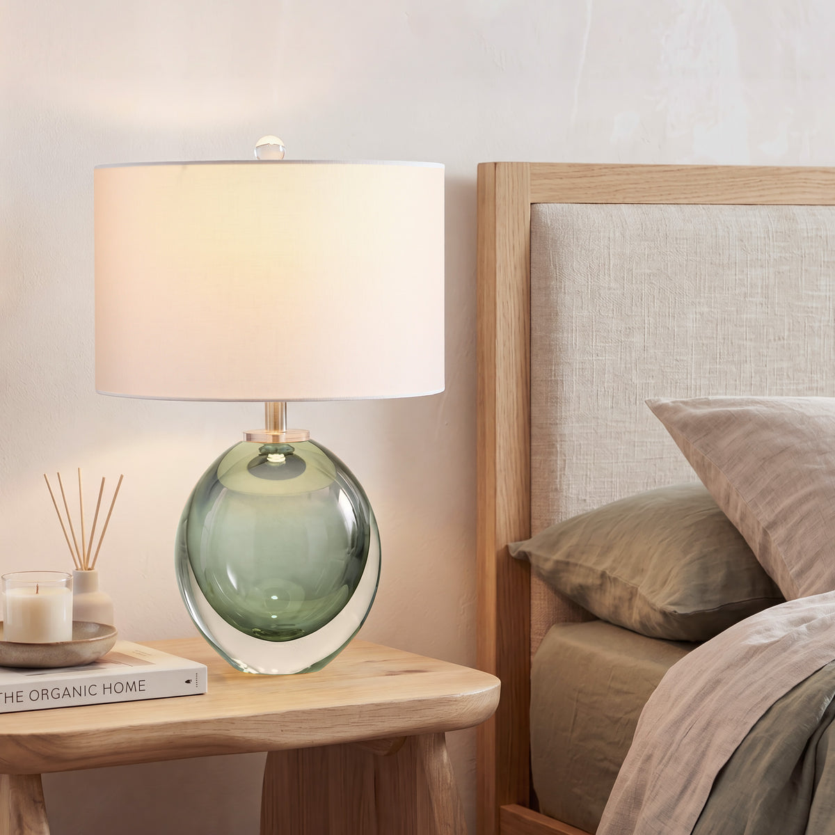 A warm glowing emerald green glass bedside lamp on a light oak nightstand next to a linen upholstered bed in a cozy organic modern bedroom