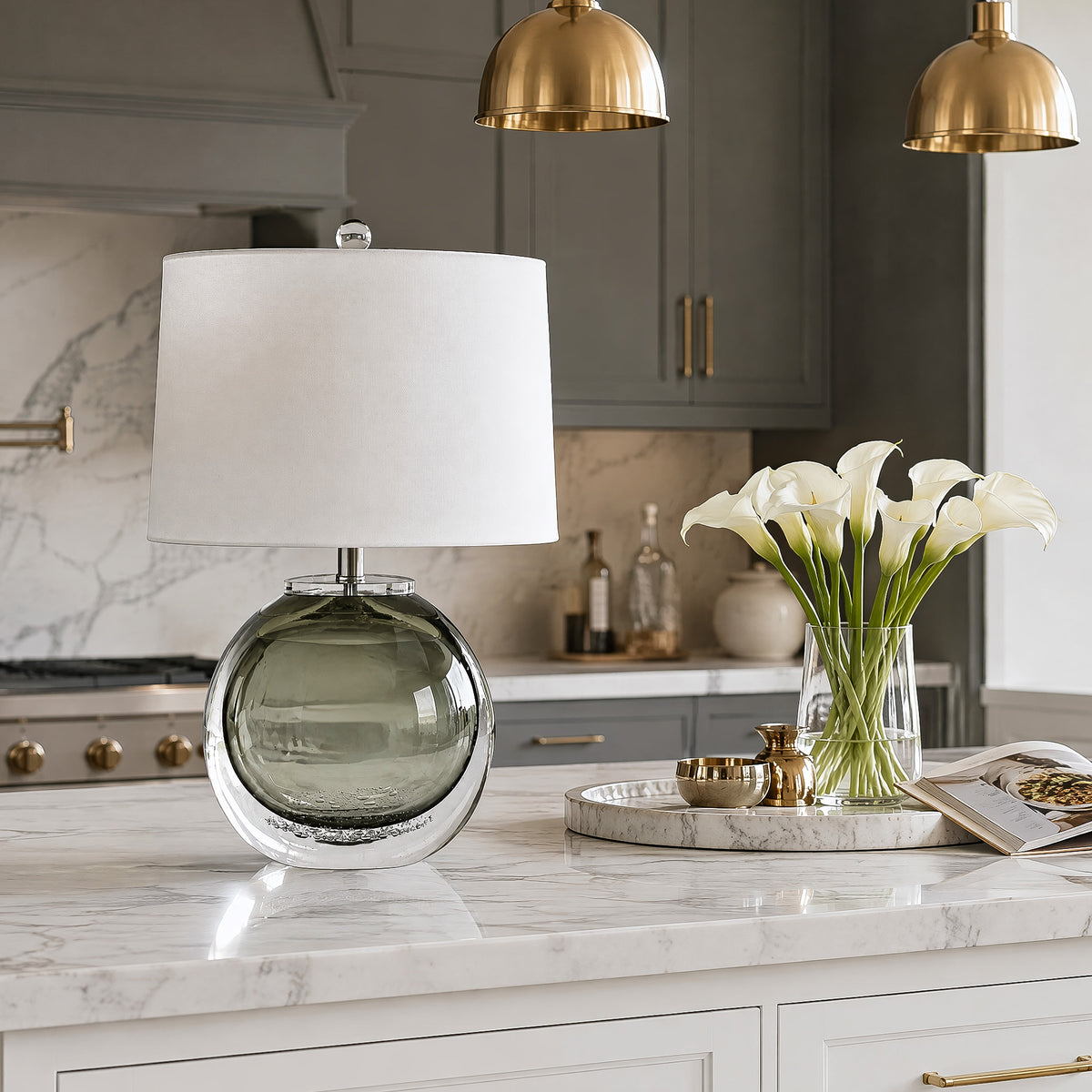 Art glass table lamp with white drum shade on a marble kitchen island near fresh flowers and golden pendant lights