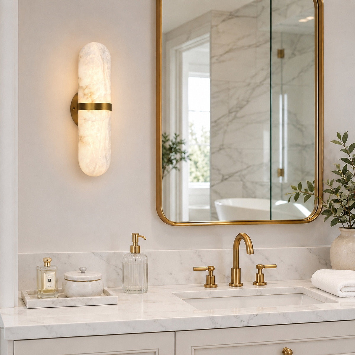 Glowing vertical capsule alabaster wall sconce mounted beside a brass frame mirror over a modern white bathroom vanity