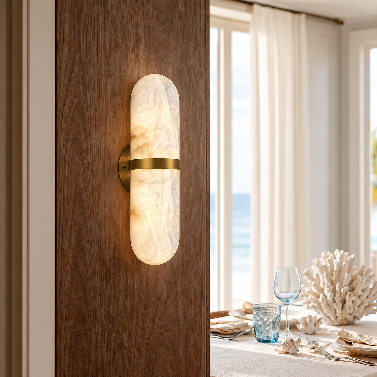 Illuminated capsule natural alabaster wall sconce mounted on dark wood wall in a bright coastal dining room setting