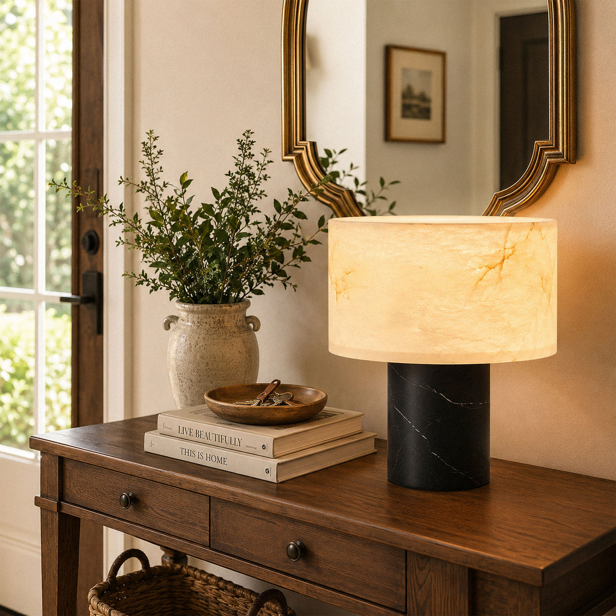 Translucent alabaster table lamp glowing on dark wooden foyer table below decorative mirror