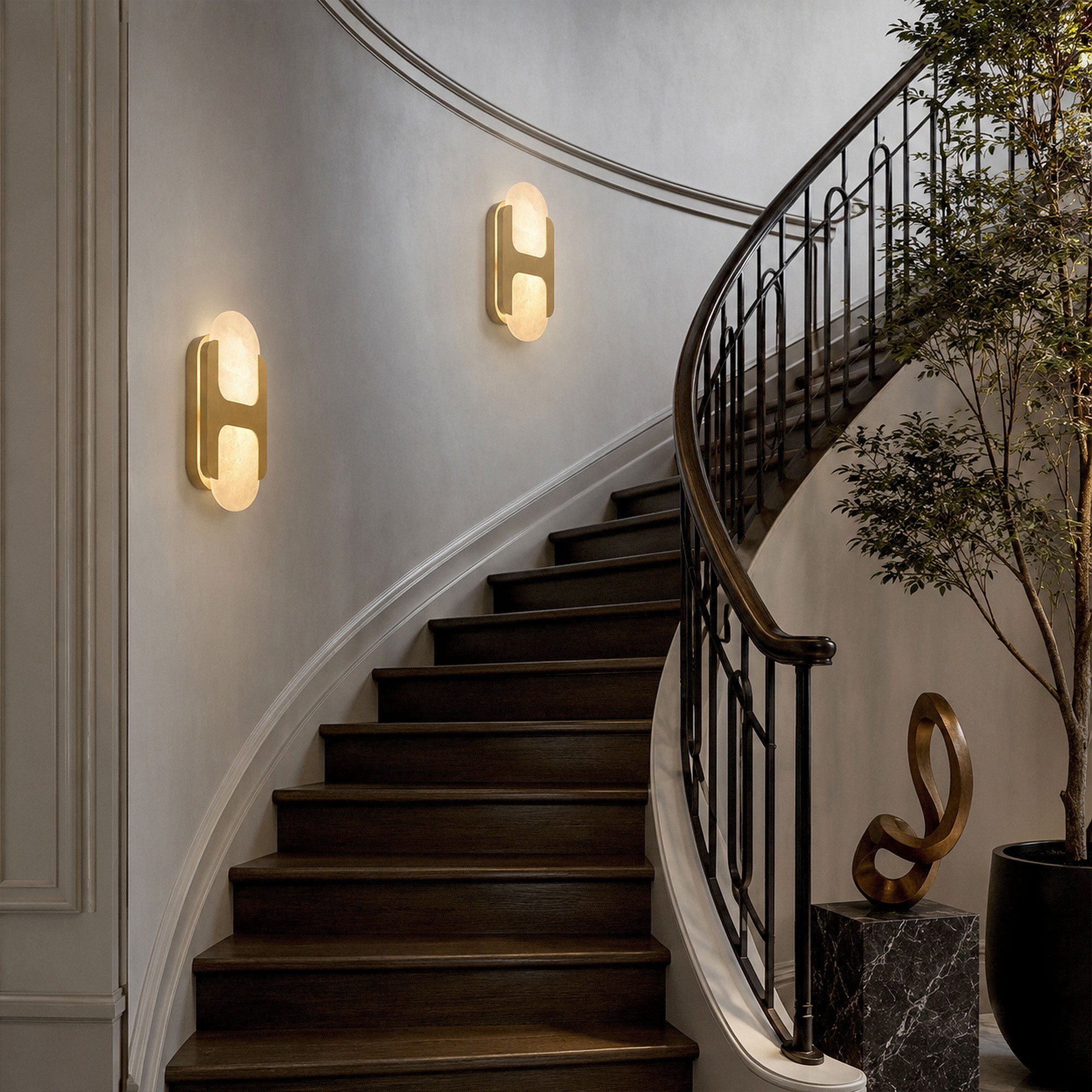 Two illuminated brass H-shaped alabaster wall sconces installed along a curved wooden staircase wall