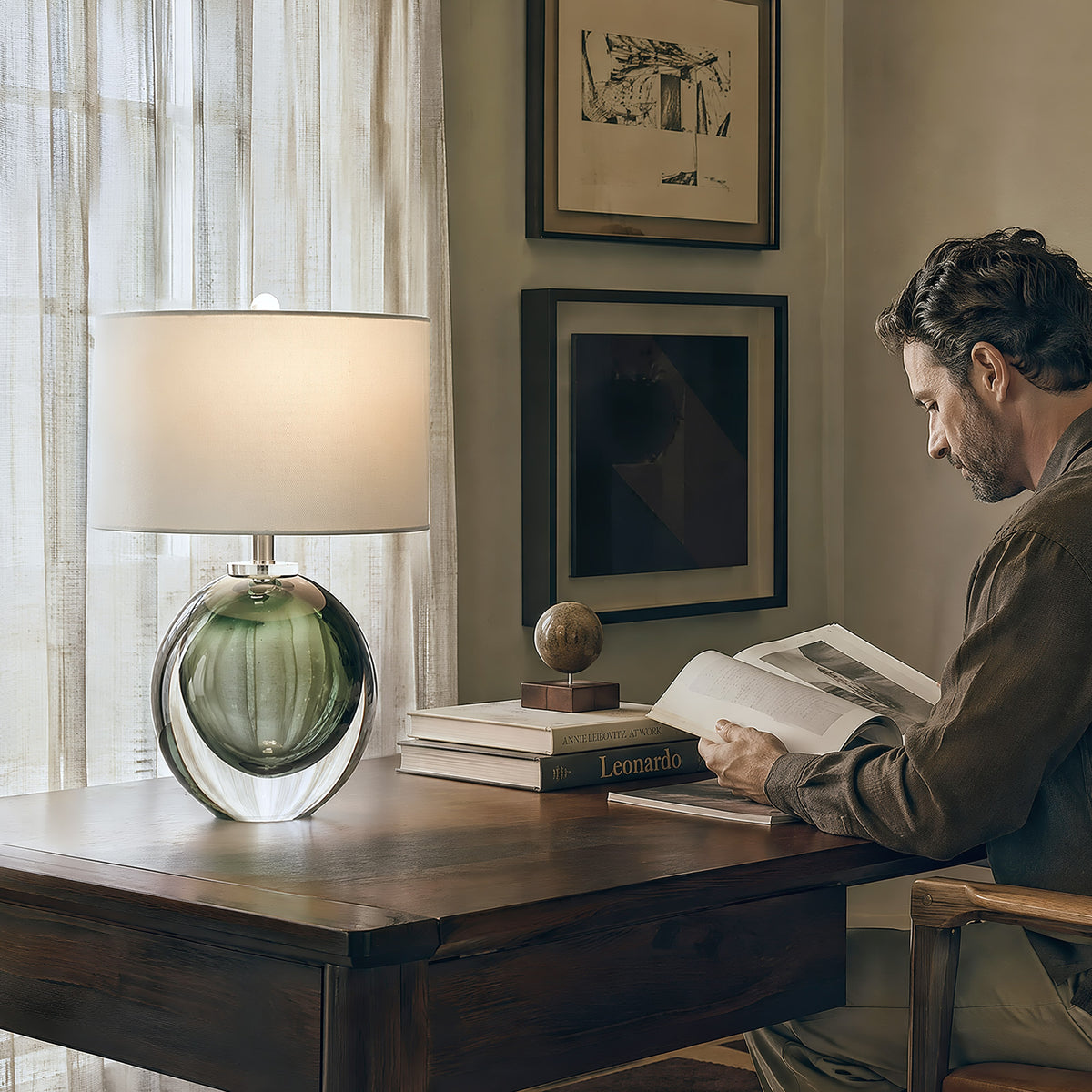 A man reading a book at a classic wooden executive desk lit by a Murano style green bubble art glass table lamp in a warm cozy home library