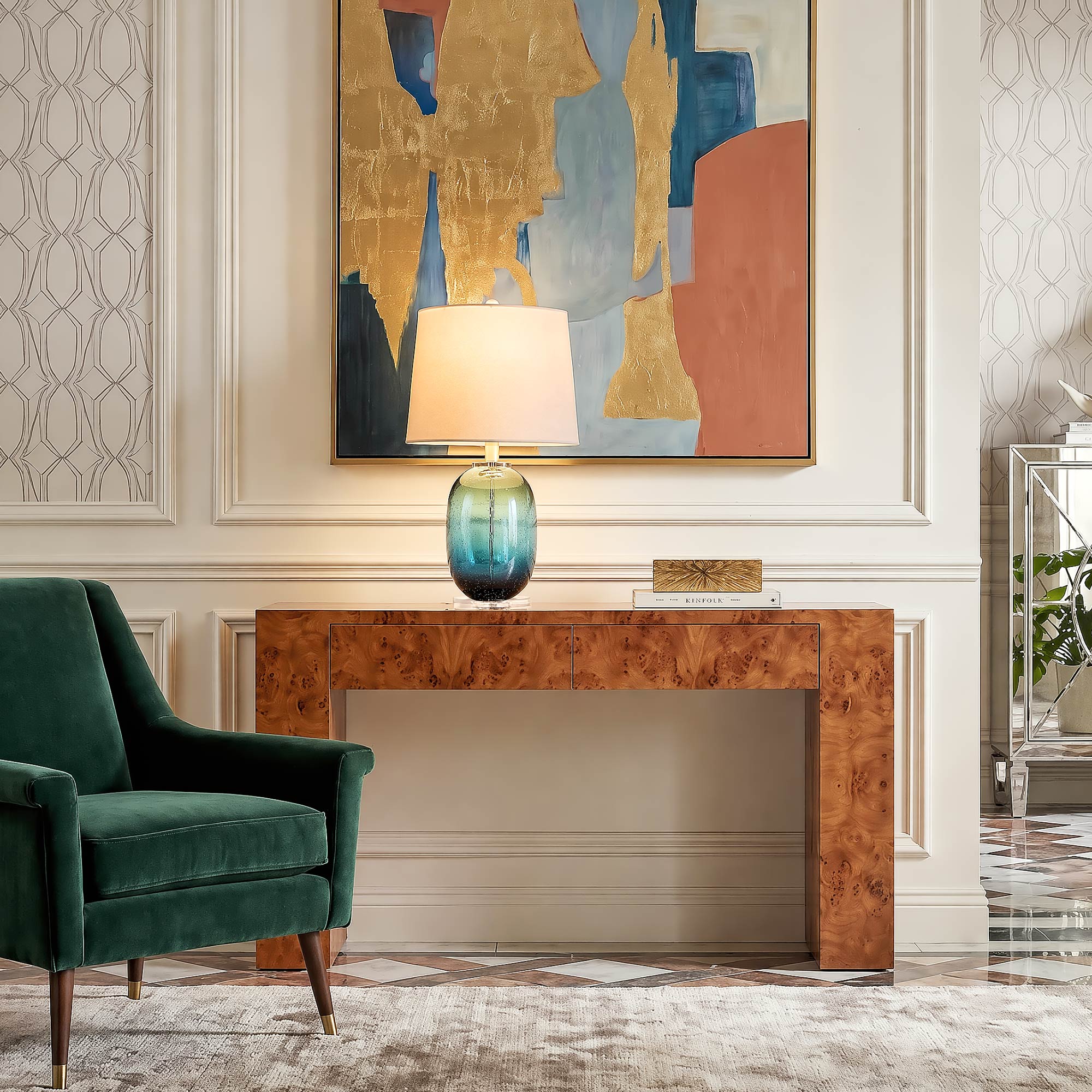 Handblown tall cylindrical blue glass lamp with floating internal bubble textures lit on a luxury burl wood entryway table under colorful abstract art