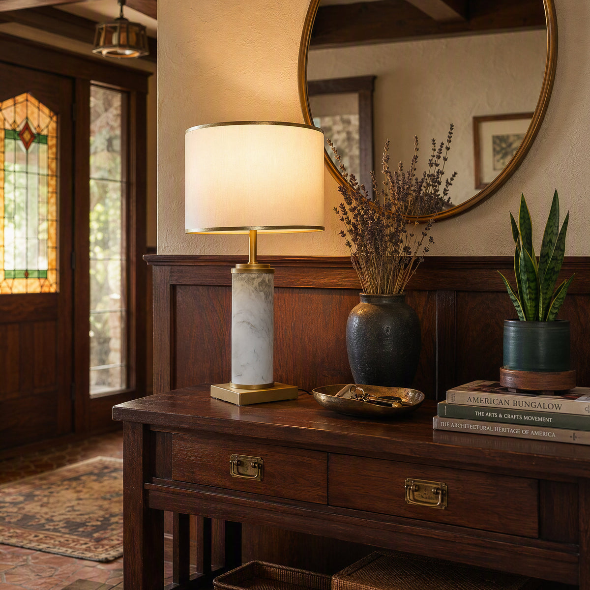 Illuminated white marble cylinder table lamp on dark wooden entry console table below round mirror