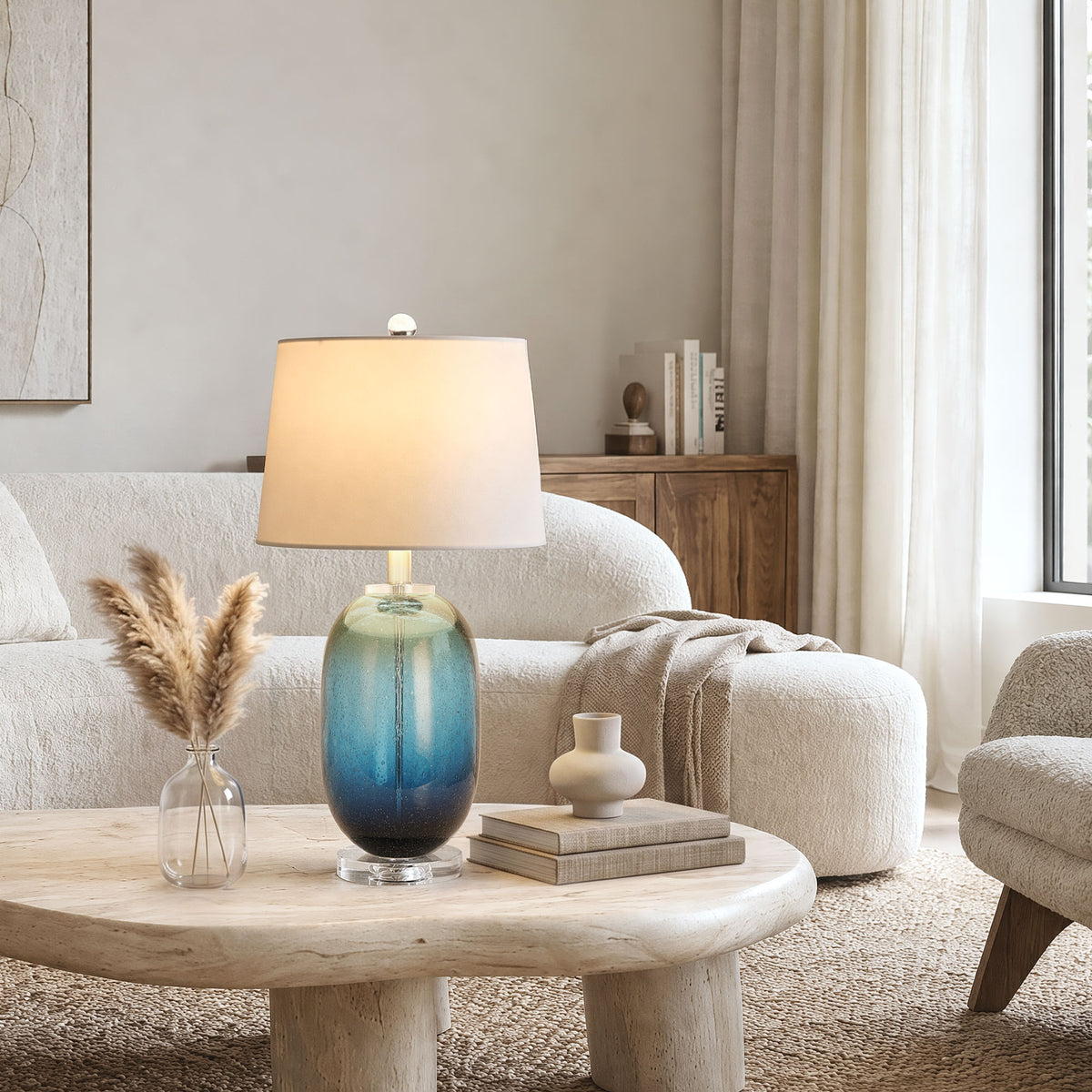 Close up view of a tall barrel shaped blue ink bubble glass table lamp illuminated on a travertine coffee table in a modern cozy cream textured living room