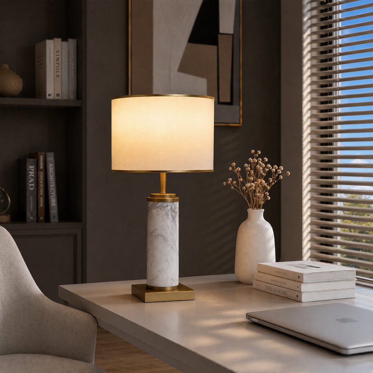 Illuminated white marble cylinder table lamp on light gray home office desk near window blinds